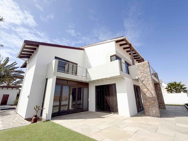 Rear view of a modern two-story house featuring stone cladding, a paved patio, and an upper-level balcony with stainless steel railings.