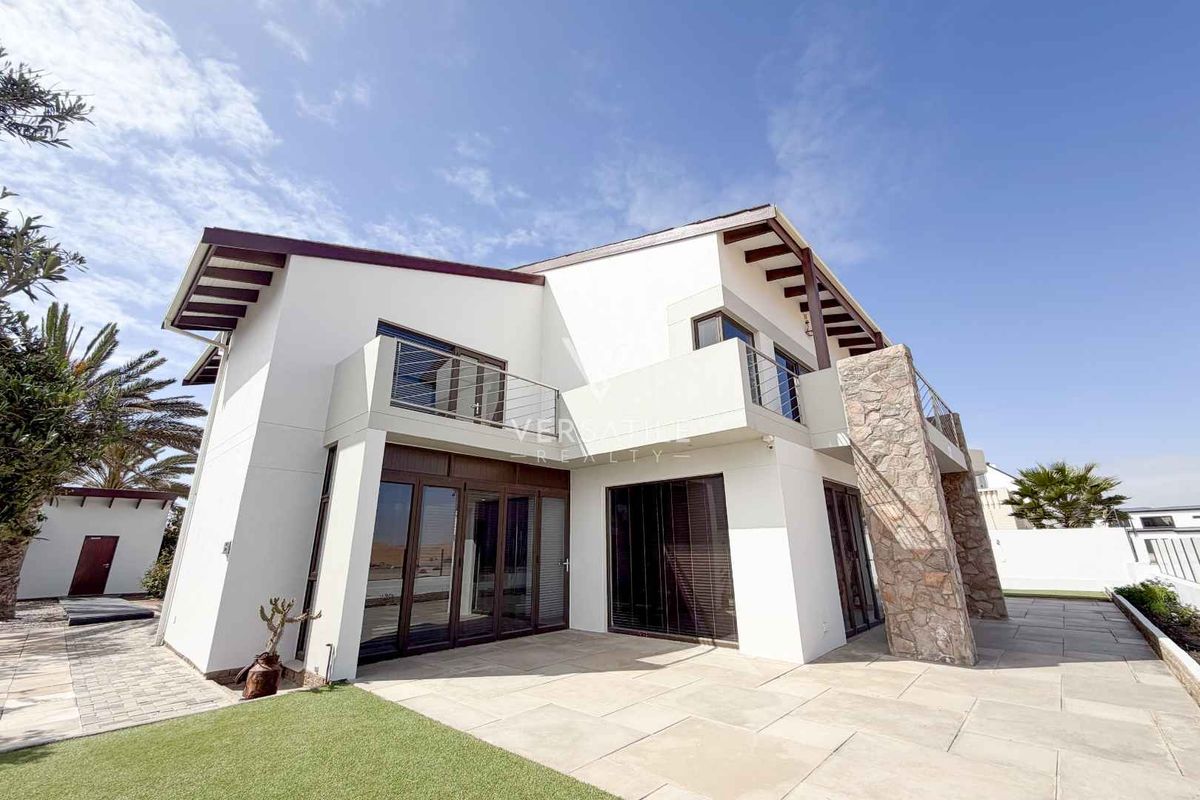 Rear view of a modern two-story house featuring stone cladding, a paved patio, and an upper-level balcony with stainless steel railings.