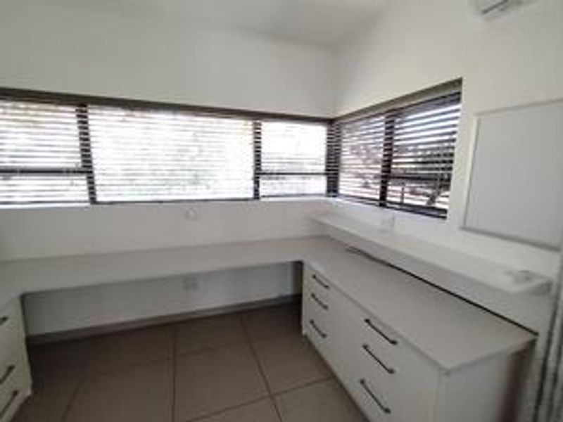 A bright, functional home office featuring extensive built-in desk space, ample cabinetry, large windows with venetian blinds, and tiled flooring.