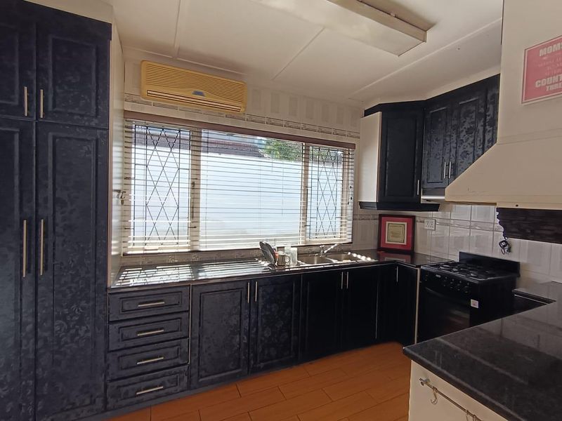 A functional kitchen featuring dark built-in cabinetry, a gas hob with extractor fan, double sink, and tiled backsplash with security bars on the window.