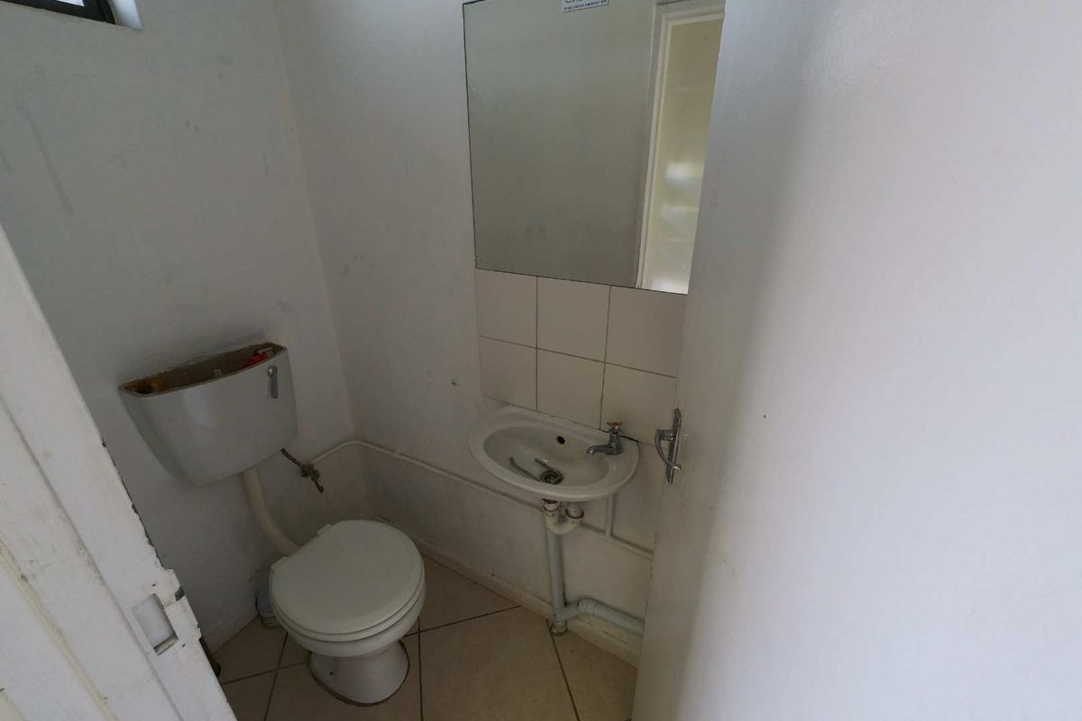 A compact industrial restroom featuring a basic toilet, wall-mounted basin, and mirror, requiring cosmetic updates and maintenance to the exposed plumbing.