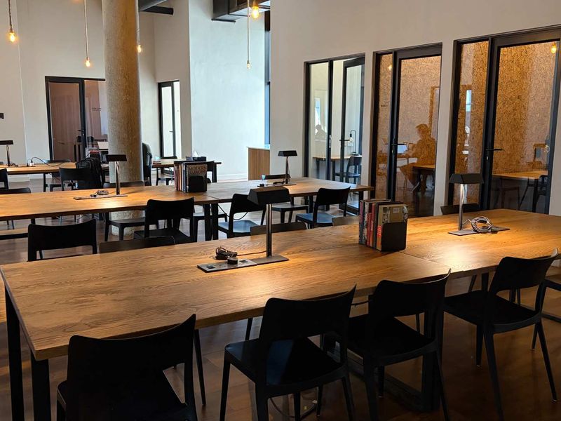 Modern open-plan office space featuring hardwood flooring, exposed concrete columns, and glass-partitioned meeting pods with contemporary lighting.