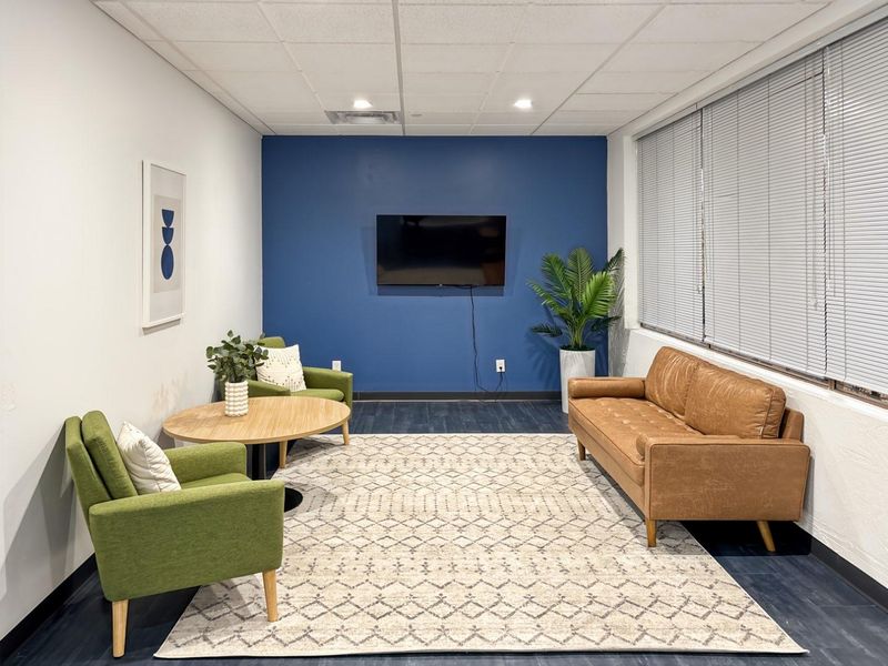 A modern office lounge area featuring a bold blue accent wall, suspended ceiling with recessed lighting, and large windows with venetian blinds.