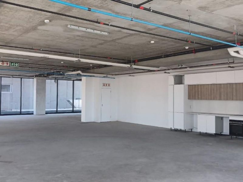 Modern open-plan office space featuring an exposed industrial ceiling, floor-to-ceiling windows for natural light, and a sleek built-in kitchenette with integrated oven.