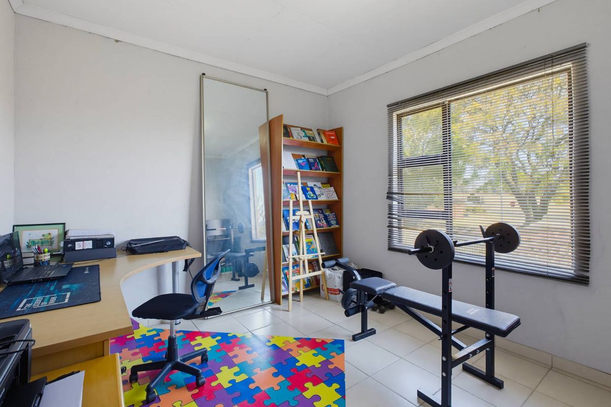 A versatile bedroom or home office space featuring tiled flooring, large window with venetian blinds, and neutral wall finishes.