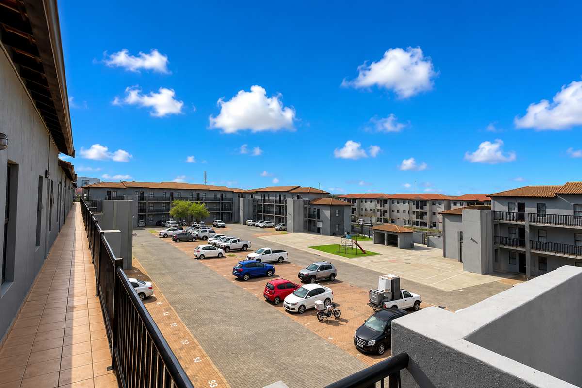 An elevated view of the apartment complex courtyard, featuring a tiled walkway, paved parking area, and multi-story residential blocks under a clear blue sky.