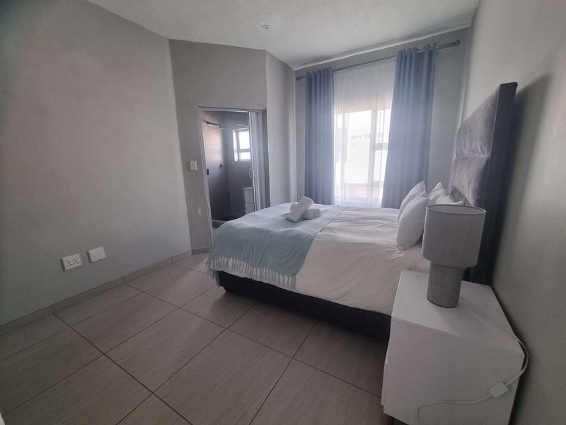 A well-maintained bedroom featuring neutral tiled flooring, an en-suite bathroom, and natural light through a window with curtains.