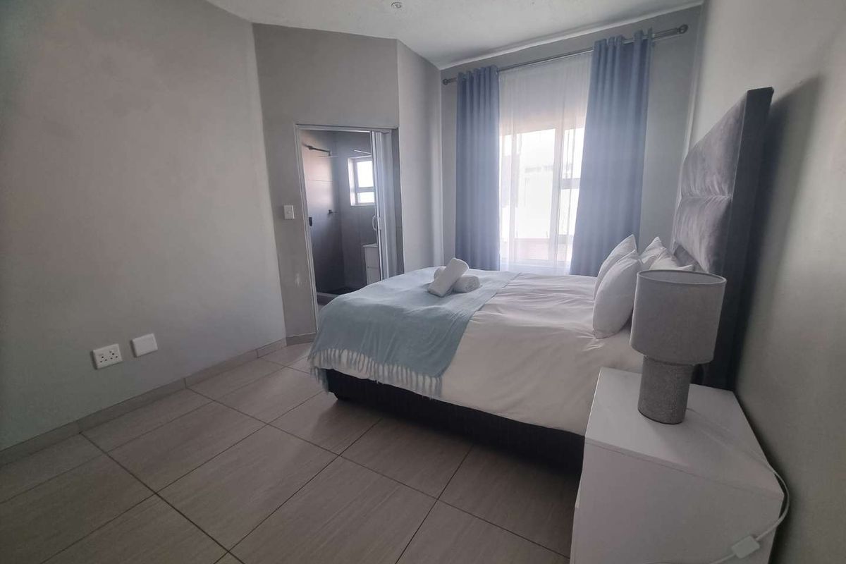 A well-maintained bedroom featuring neutral tiled flooring, an en-suite bathroom, and natural light through a window with curtains.