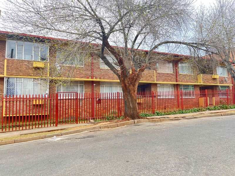 Front exterior view of a multi-story brick apartment building featuring metal security fencing and mature trees along the street frontage.
