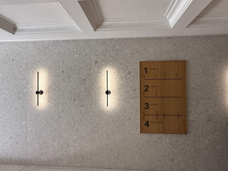 A modern office hallway featuring elegant terrazzo wall cladding, recessed coffered ceiling details, and minimalist wall-mounted lighting fixtures with a professional directory board.