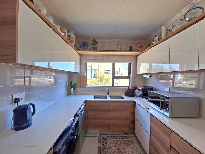 Modern galley-style kitchen featuring sleek white and wood-grain cabinetry, stone countertops, a double bowl sink, and integrated appliances.