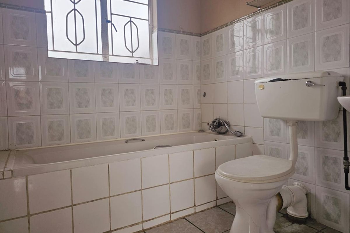 A dated bathroom featuring a built-in bathtub, standard toilet, and tiled walls with a security-barred window.