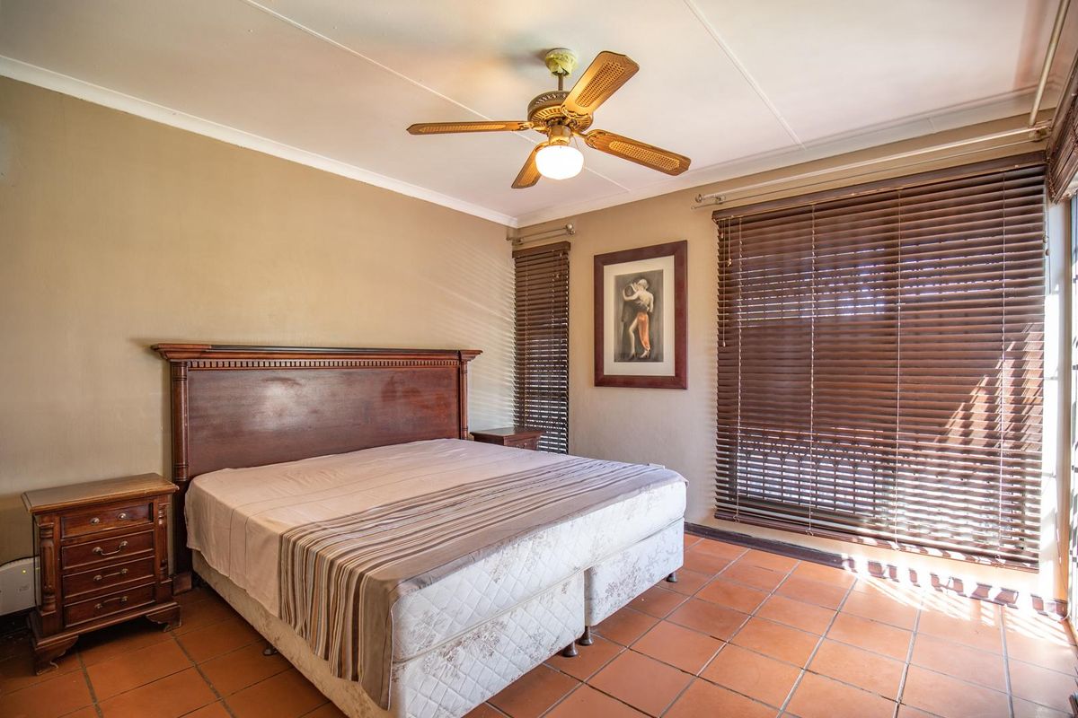 A spacious bedroom featuring terracotta floor tiles, a ceiling fan, and large windows fitted with wooden venetian blinds.
