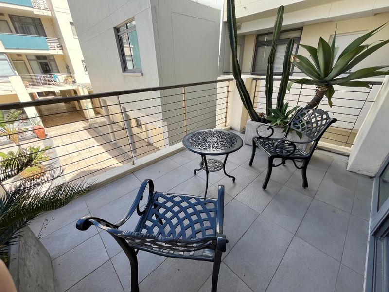 A private balcony featuring tiled flooring and a metal railing, overlooking the apartment complex courtyard.