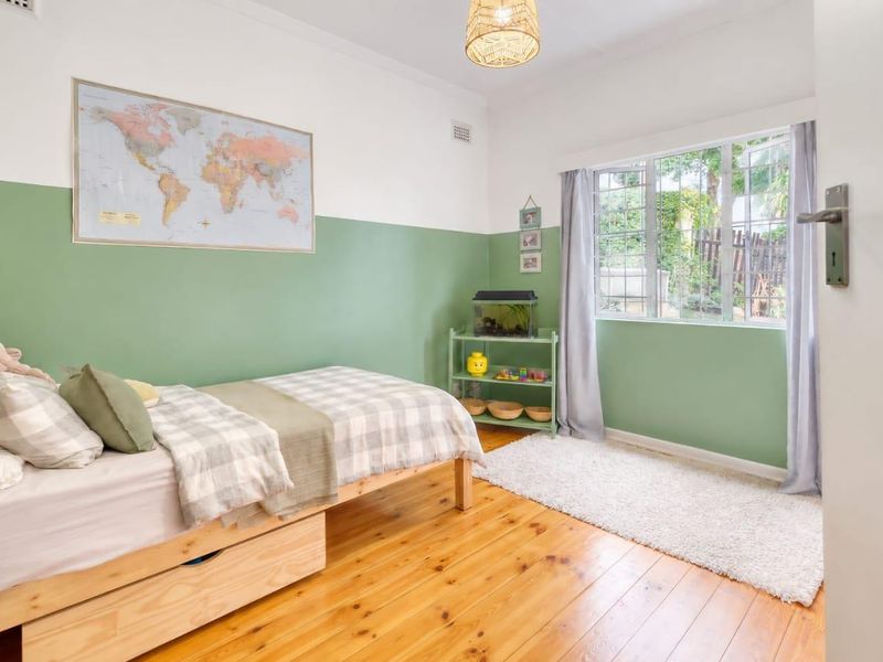 A bright bedroom featuring polished timber flooring, two-tone wall paint, and a window with security bars overlooking the garden.