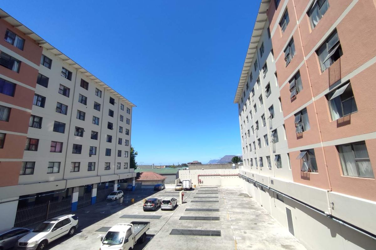 Exterior view of an apartment complex featuring multi-story residential blocks with a central paved parking area and clear sky.