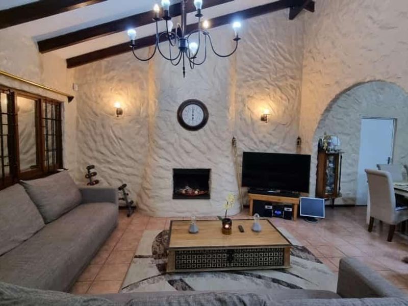 A cozy living room featuring textured plaster walls, exposed timber ceiling beams, a built-in fireplace, and an arched entryway leading to the dining area.