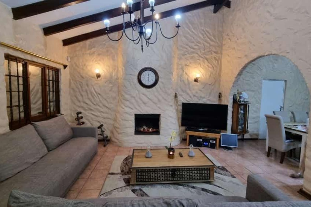 A cozy living room featuring textured plaster walls, exposed timber ceiling beams, a built-in fireplace, and an arched entryway leading to the dining area.
