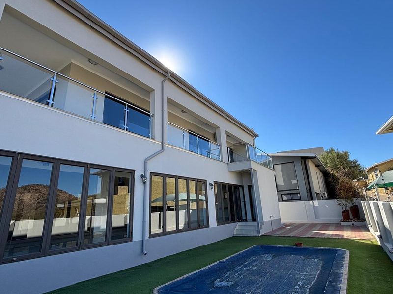 Rear view of a modern two-story house featuring a swimming pool, artificial lawn, paved patio, and upper-level balconies with glass balustrades.