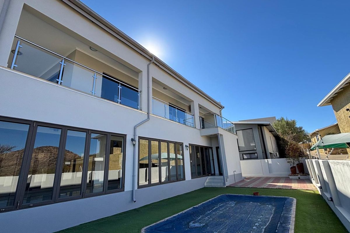 Rear view of a modern two-story house featuring a swimming pool, artificial lawn, paved patio, and upper-level balconies with glass balustrades.