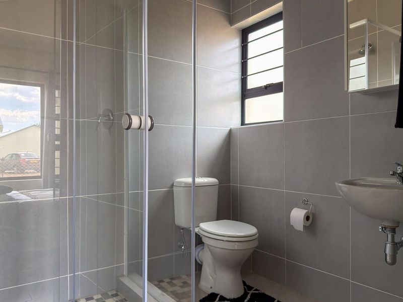 Modern bathroom featuring grey ceramic wall tiles, a glass shower enclosure, a wall-mounted basin, and a toilet with a louvered window for ventilation.