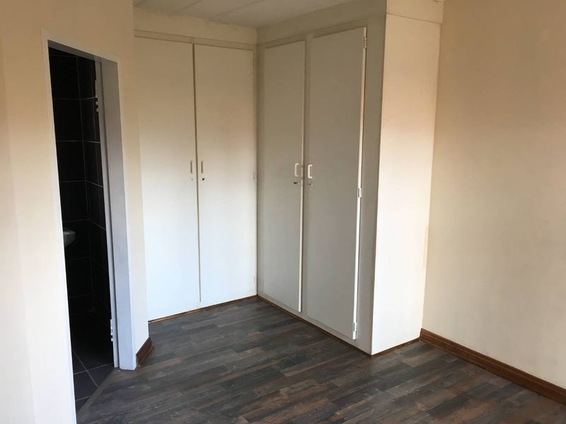 A clean, empty bedroom featuring built-in cupboards and modern laminate flooring, with an adjacent doorway leading to a tiled bathroom area.