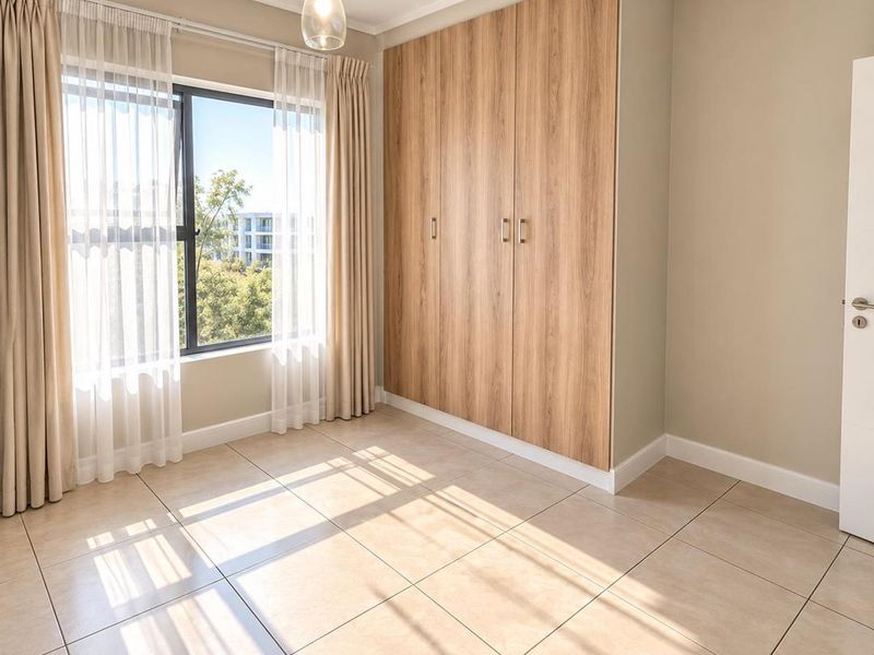 A bright, empty bedroom featuring neutral tiled flooring, a large built-in wooden wardrobe, and a window providing natural light.