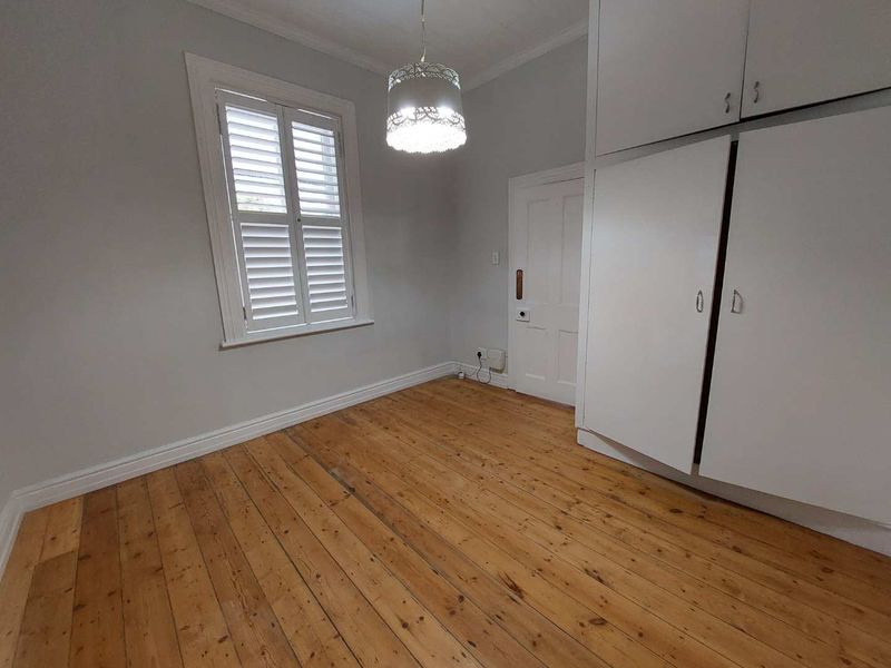 A bright, empty bedroom featuring original timber flooring, built-in storage cupboards, and plantation shutters on the window.