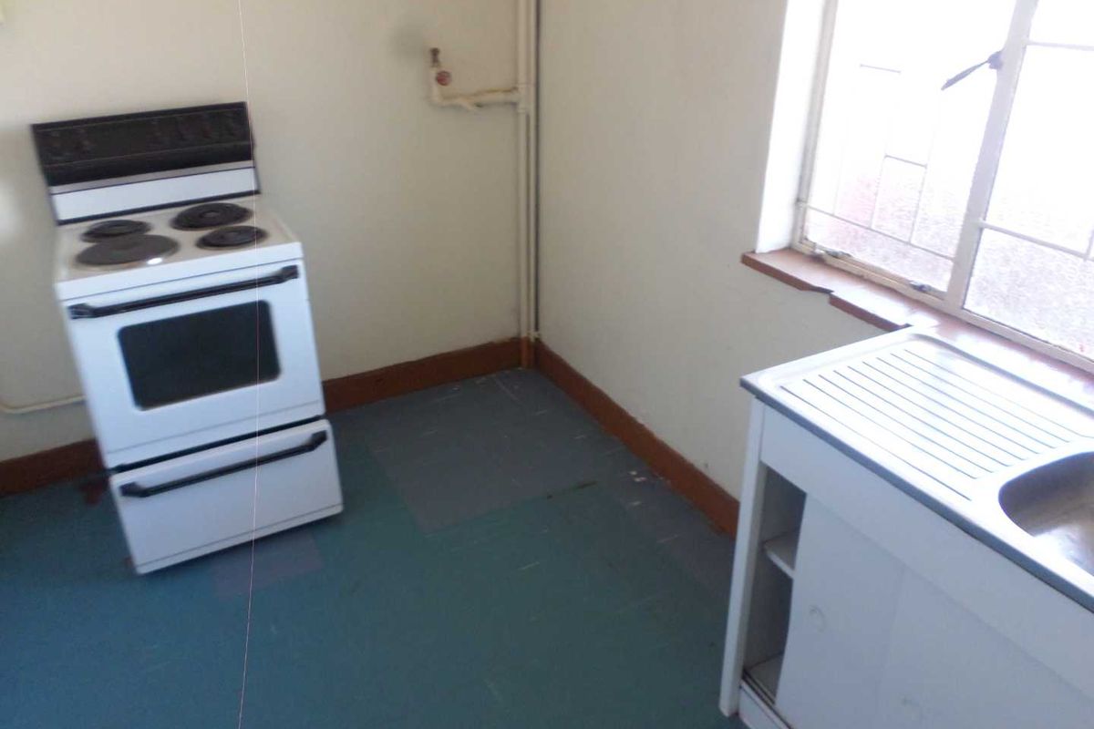 A basic, dated kitchen area featuring a freestanding stove, a single sink unit, and a window with security bars.
