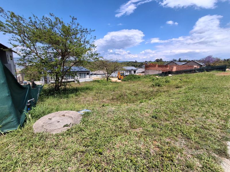A view of a vacant residential plot featuring natural grass cover, a manhole access point, and surrounding residential development in the background.