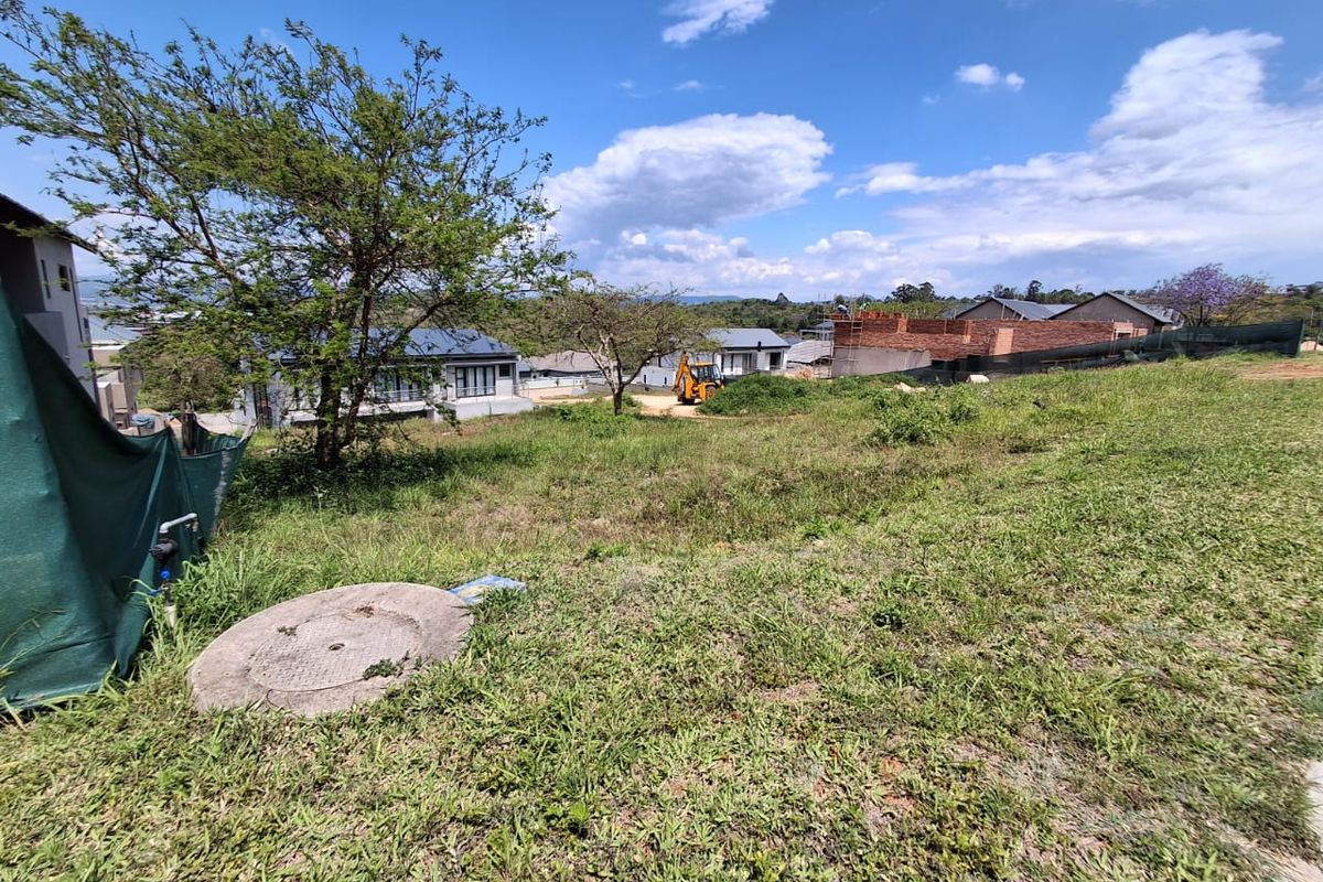 A view of a vacant residential plot featuring natural grass cover, a manhole access point, and surrounding residential development in the background.