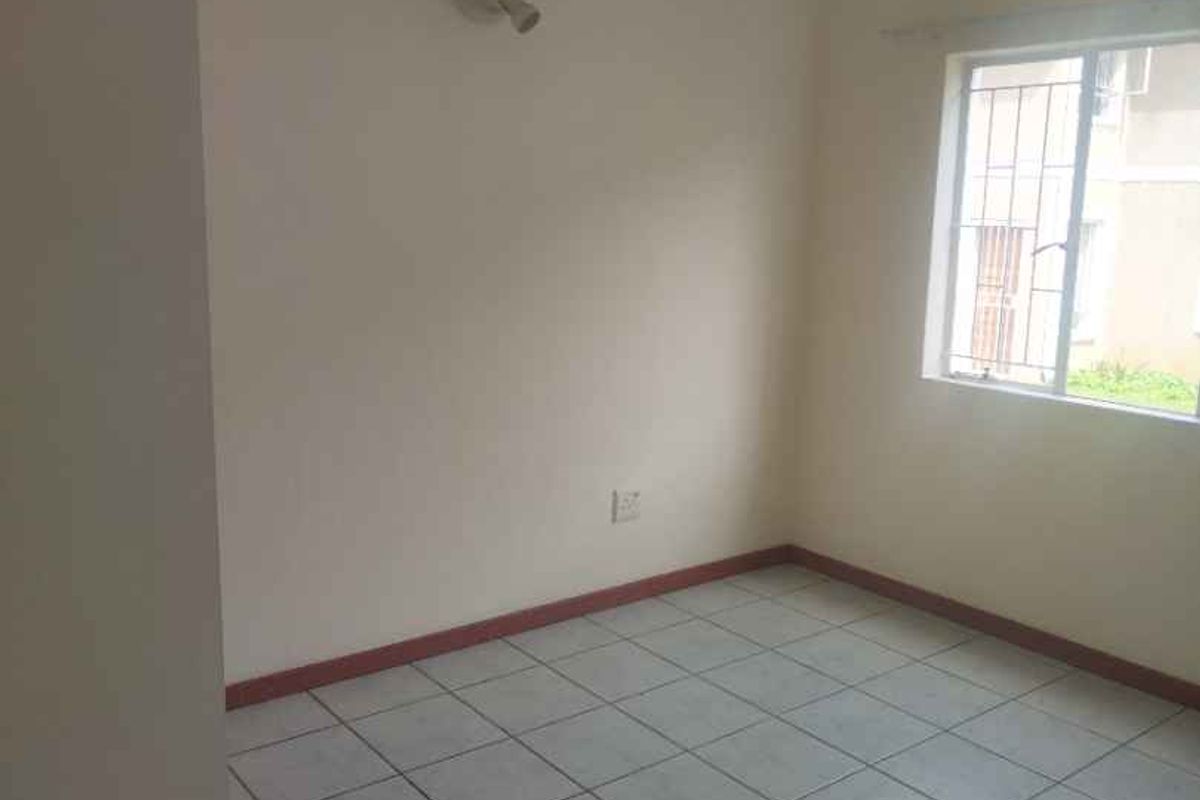 A vacant bedroom featuring neutral tiled flooring, a single wall-mounted light fixture, and a window with security bars.