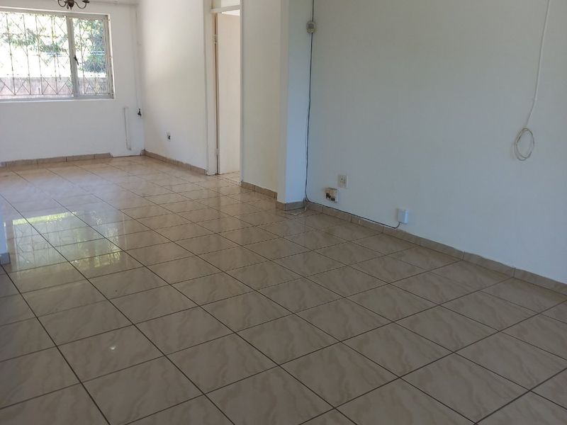 An empty, open-plan living area featuring neutral ceramic floor tiles, an arched transition to an adjacent room, and a window with security bars.