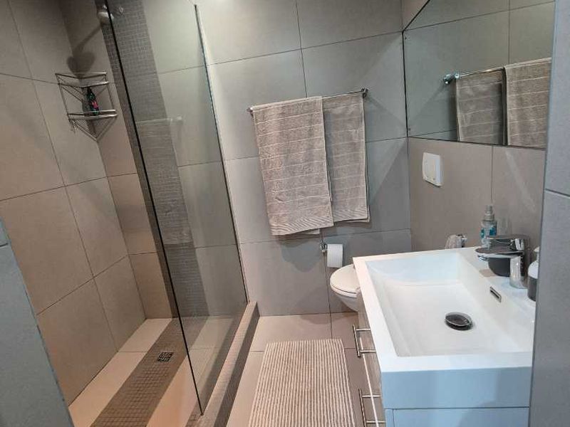 Modern bathroom featuring a glass-enclosed walk-in shower, wall-mounted vanity with basin, large mirror, and contemporary grey tiling throughout.