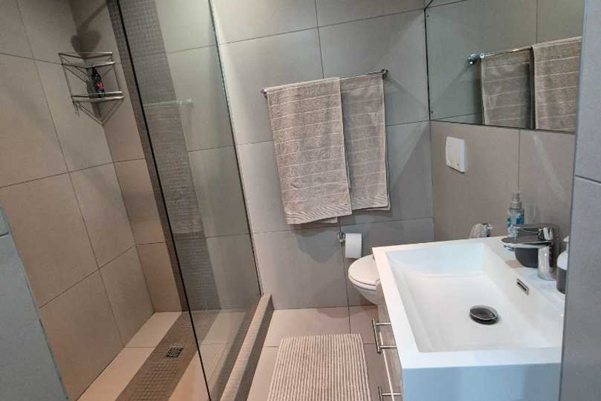 Modern bathroom featuring a glass-enclosed walk-in shower, wall-mounted vanity with basin, large mirror, and contemporary grey tiling throughout.