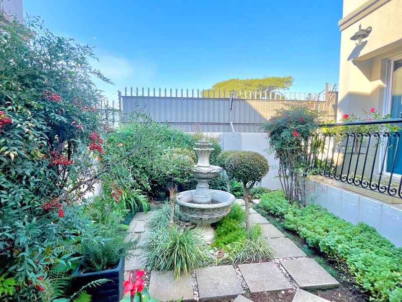 A well-maintained garden area featuring a central stone fountain, paved pathways, and established perimeter fencing.