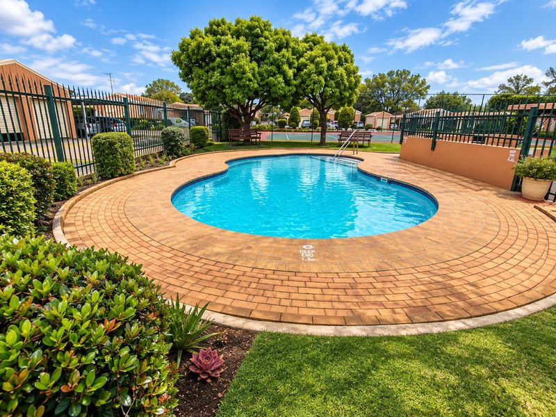A well-maintained communal swimming pool area featuring brick paving, landscaped gardens, and secure metal perimeter fencing within the townhouse complex.