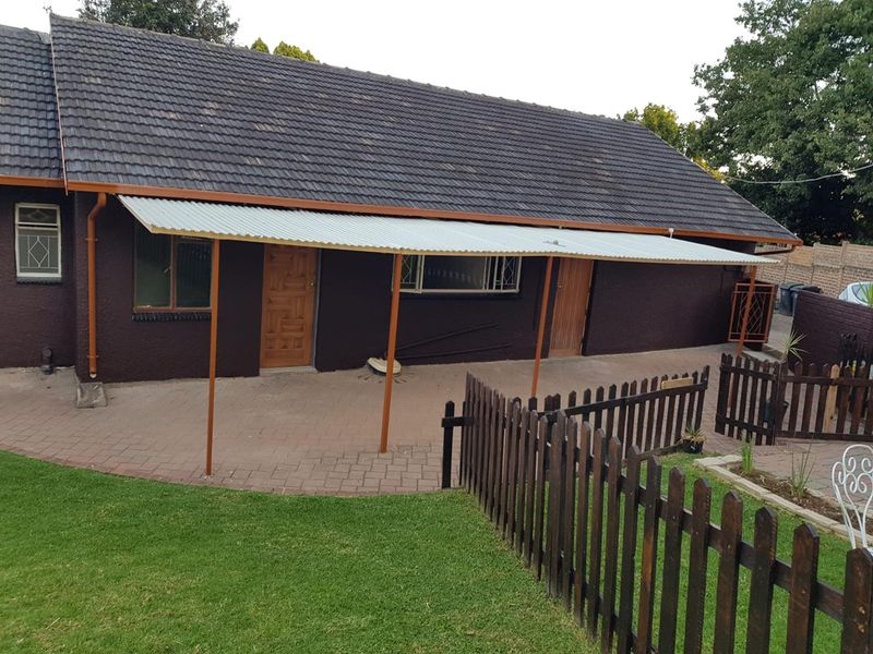 Rear view of the property featuring a paved patio area covered by a corrugated metal roof, enclosed by a timber picket fence and lawn.
