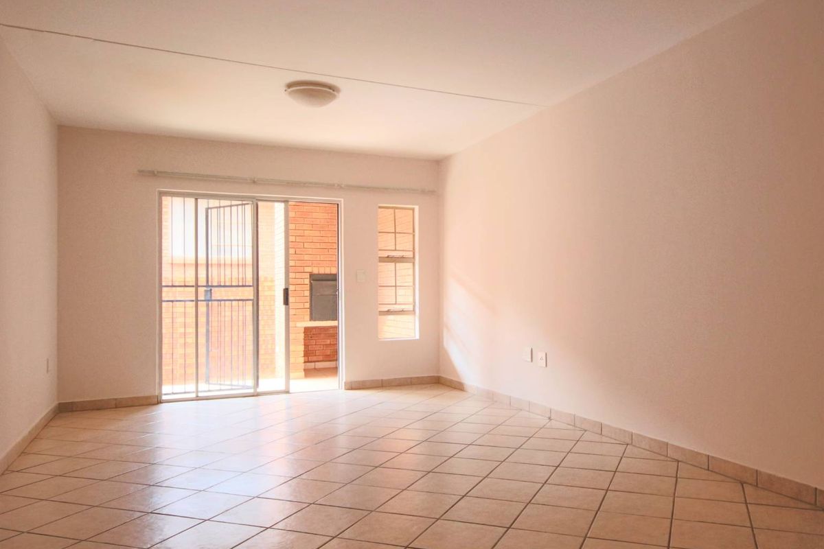 An empty, sunlit living room featuring neutral tiled flooring and a sliding glass door providing access to an outdoor patio area.