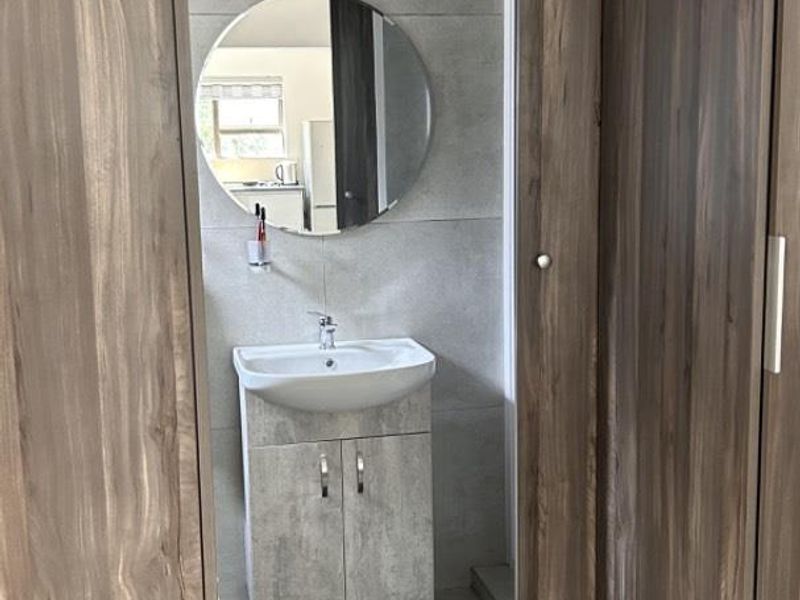 A modern bathroom featuring a vanity cabinet with a ceramic basin, round mirror, and large format grey wall tiles, framed by built-in wooden cupboards.