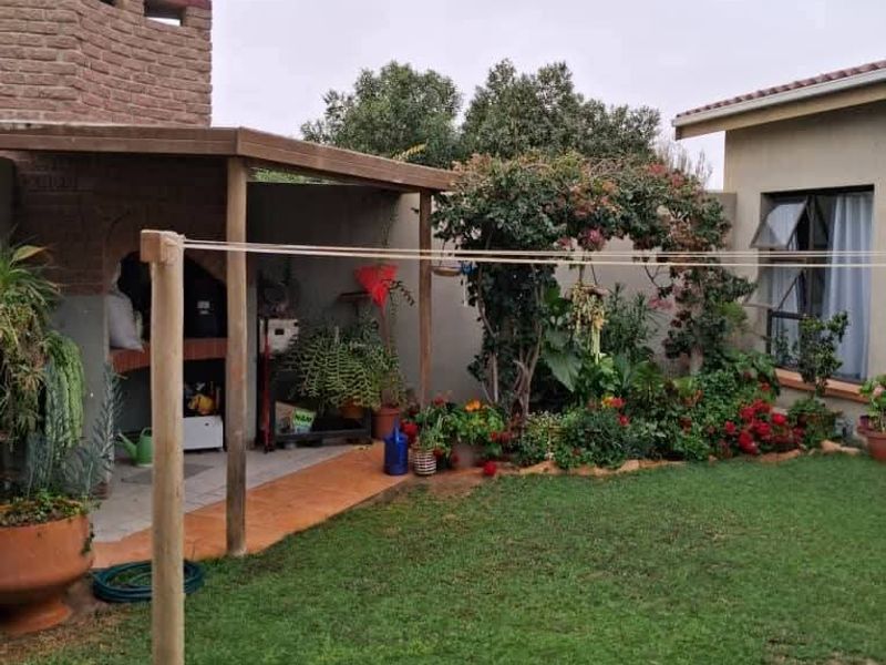 A rear garden area featuring a lawn, a covered patio with a built-in braai, and a brick chimney structure.