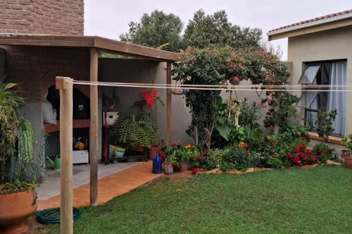 A rear garden area featuring a lawn, a covered patio with a built-in braai, and a brick chimney structure.