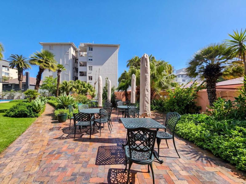 A well-maintained outdoor patio area featuring brick paving, surrounded by lush landscaped gardens and a boundary wall, ideal for communal relaxation.