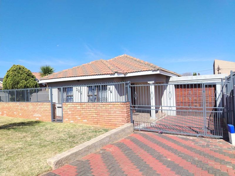 Front elevation of a single-story house featuring a tiled roof, brick boundary wall, steel security fencing, and a paved driveway with a garage.