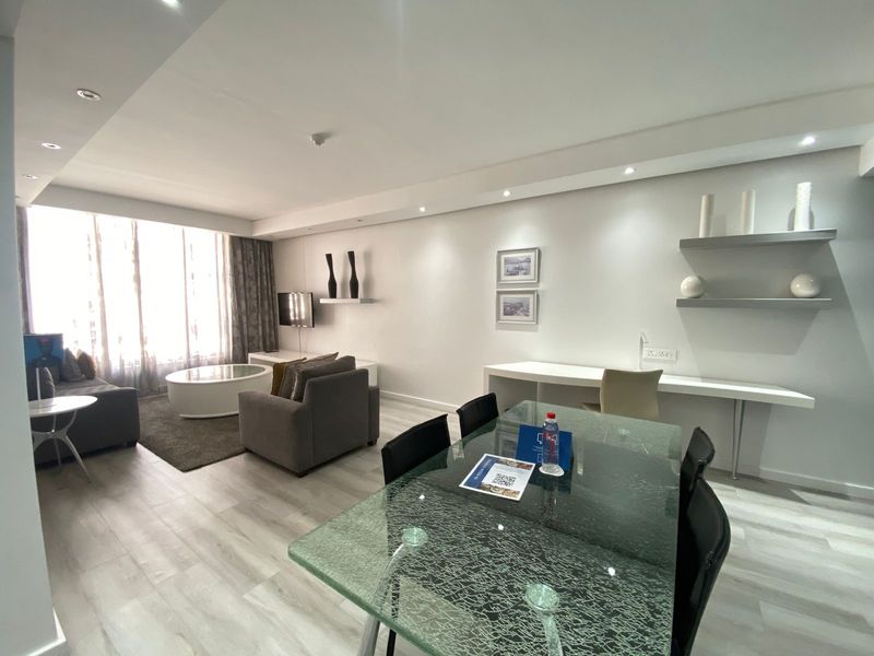 A modern open-plan living and dining area featuring light laminate flooring, recessed lighting, a built-in desk, and floating shelving.