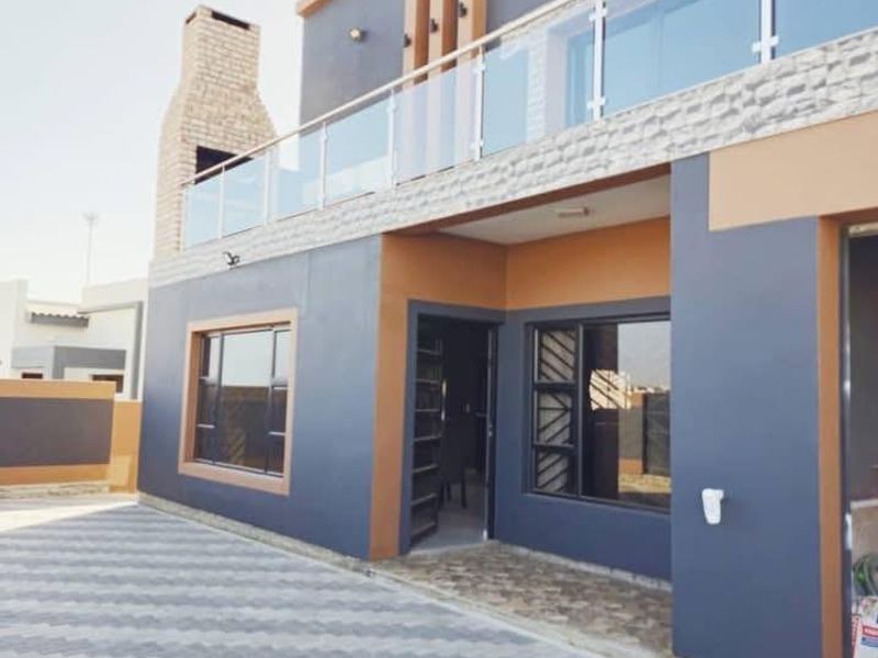 Modern rear exterior view featuring a paved patio area, stone-clad outdoor fireplace, and a balcony with a contemporary glass balustrade.