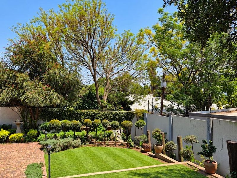 A well-maintained private garden featuring manicured lawn, brick paved pathways, established trees, and secure perimeter walling with electric fencing.