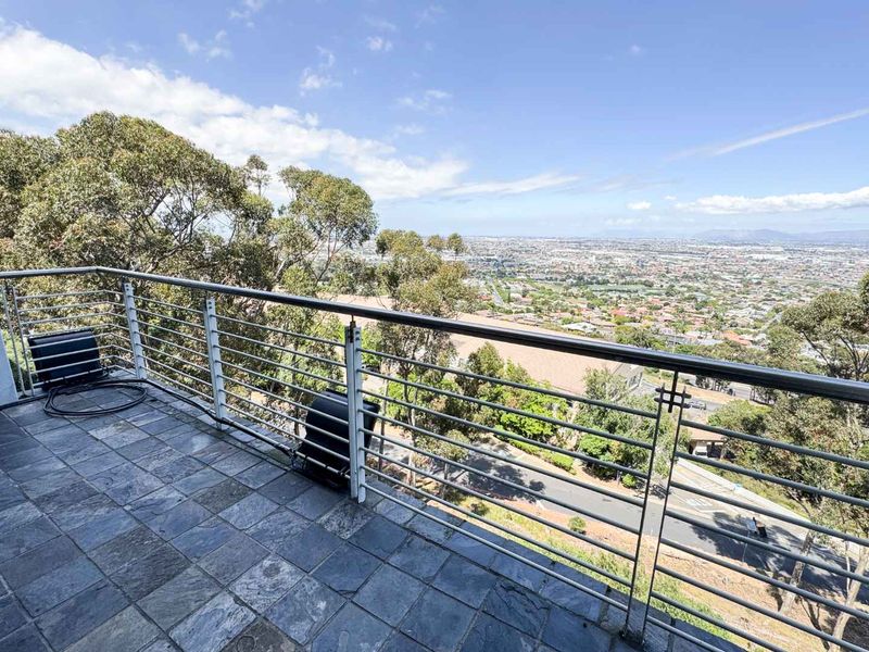 A spacious balcony featuring stone tile flooring and a modern stainless steel railing, offering expansive elevated views of the surrounding landscape and city.