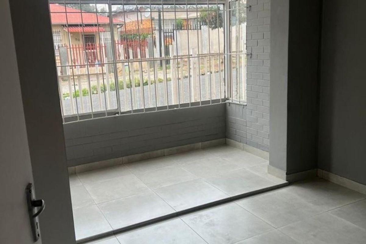 A bright, empty interior space featuring large windows with security bars, neutral tiled flooring, and painted brick accents.