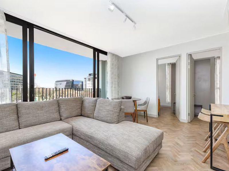 Bright open-plan living area featuring herringbone timber flooring, track lighting, and large sliding glass doors opening onto a private balcony with urban views.